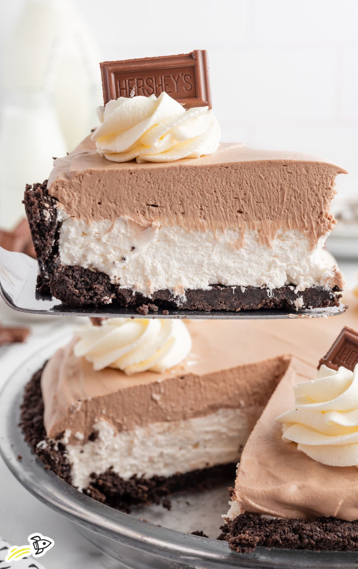 A close-up of a slice of no-bake Hershey pie with a chocolate cookie crust, creamy white filling, and fluffy chocolate mousse topping, garnished with a swirl of whipped topping and a piece of Hershey’s chocolate bar