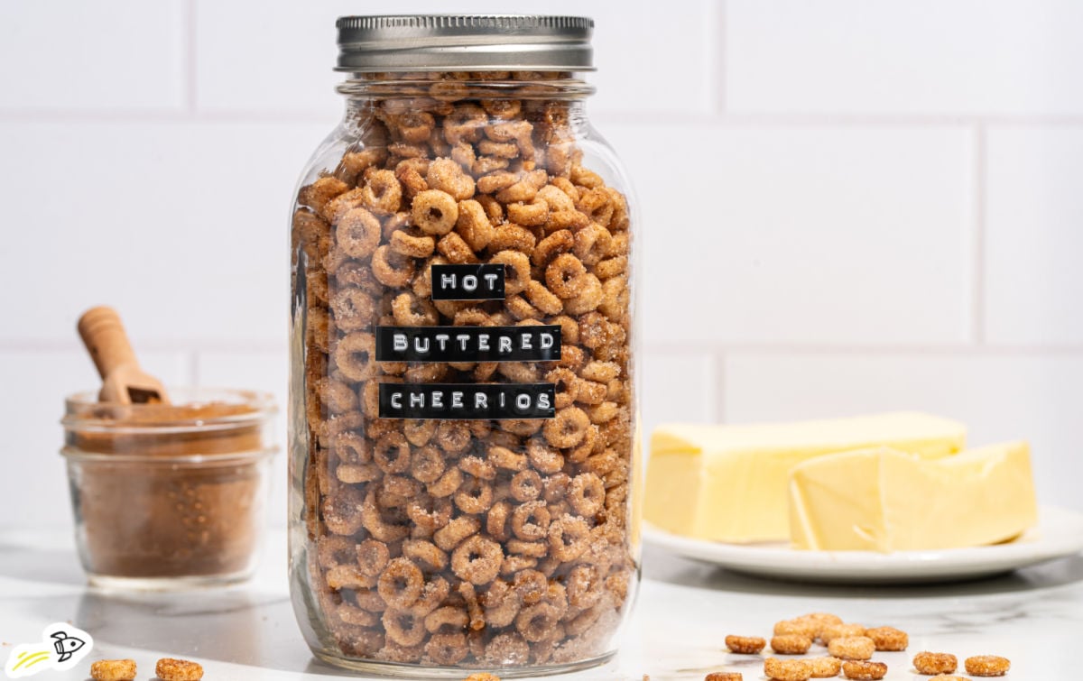 Glass jar filled with hot buttered Cheerios labeled with black text strips, surrounded by butter on a plate and a small jar of cinnamon sugar in the background.