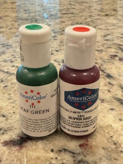 Two bottles of AmeriColor gel food coloring, Leaf Green and Super Red, side by side on a granite countertop.