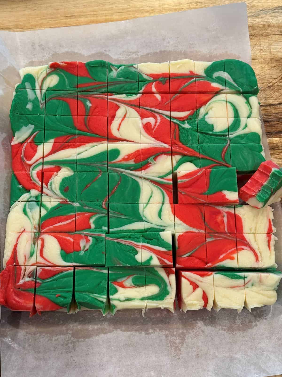 Squares of red, green and white swirled Christmas fudge on parchment paper, with one piece pulled slightly away from the rest.