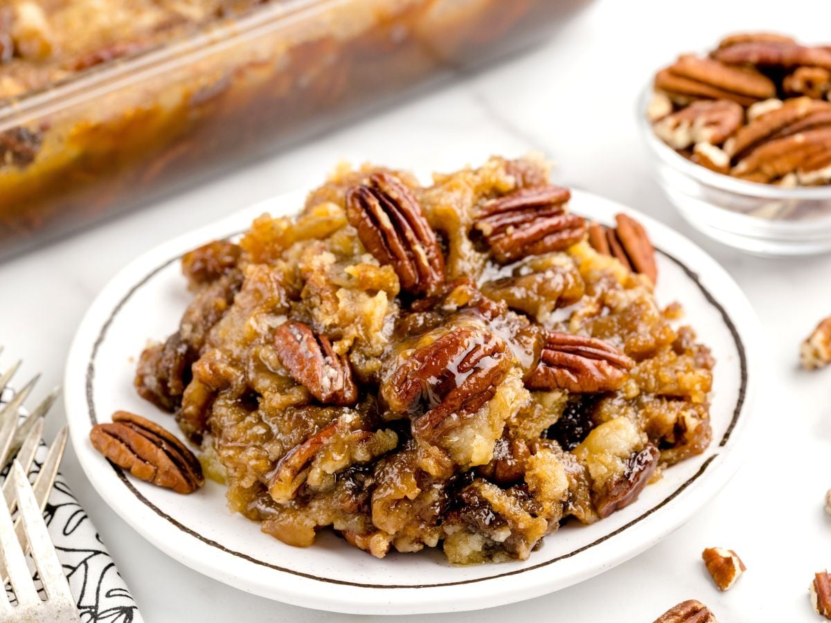 A serving of gooey Pecan Dump Cake casserole on a white plate, topped with whole pecans. In the background, there’s a casserole dish and bowl of pecans, with forks and scattered pecans nearby.