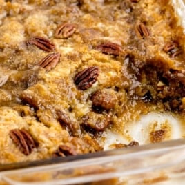 A close-up of a partially served Pecan Dump Cake in a glass baking dish, featuring a golden-brown, gooey surface topped with whole pecans and a caramelized texture.
