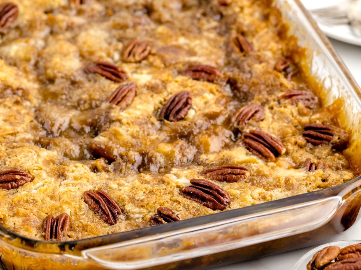 A close-up of Pecan Dump Cake in a glass dish, topped with pecan halves and a golden-brown, crumbly layer, suggesting a sweet, gooey texture.
