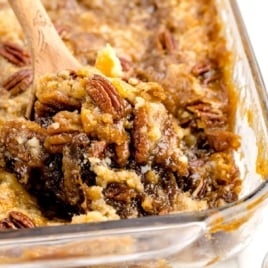 Close-up of a gooey pecan pie cobbler, similar to a Pecan Dump Cake, in a glass baking dish. A wooden spoon scoops out the dessert, revealing its golden, crumbly topping and plenty of visible pecan halves.