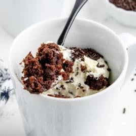 A spoon scoops a rich Mug Cake with Cake Mix, topped with whipped cream and chocolate sprinkles, from a white mug.
