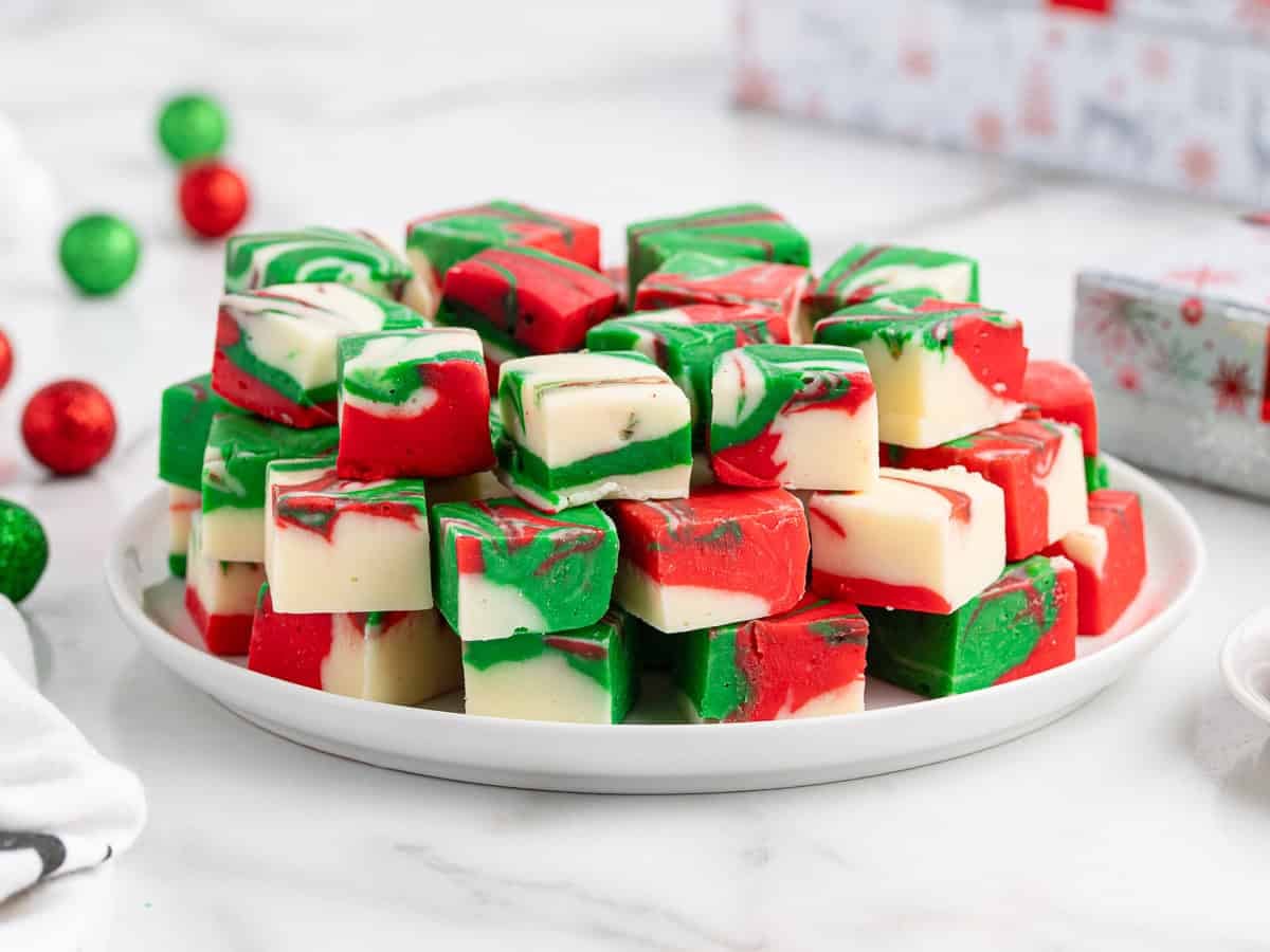 A plate filled with cubes of red, green, and white swirled holiday fudge, perfect for any Christmas Fudge Recipe, rests on a marble surface. Christmas ornaments and a wrapped gift are in the background.