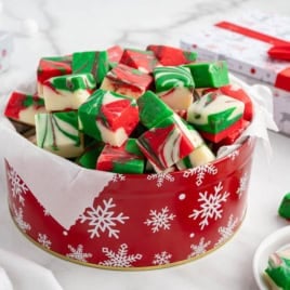 A red tin with white snowflakes holds festive red, green, and white swirled fudge squares made from a classic Christmas Fudge Recipe. Wrapped gifts and holiday decorations are in the background on a marble surface.