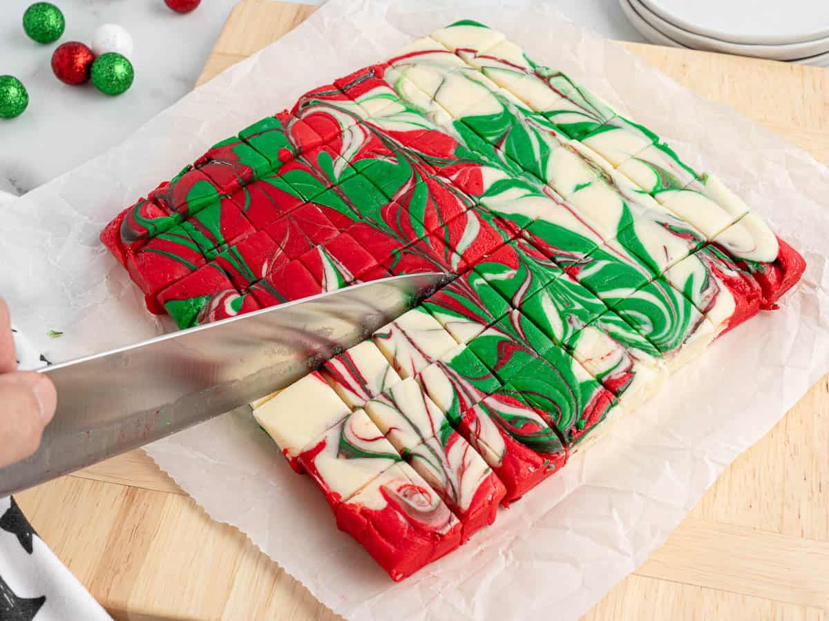 A hand is slicing festive swirled Christmas Fudge with red, green, and white colors on a parchment-lined cutting board. Decorative ornaments and stacked plates are visible in the background, setting the perfect scene for this delightful Christmas Fudge Recipe.