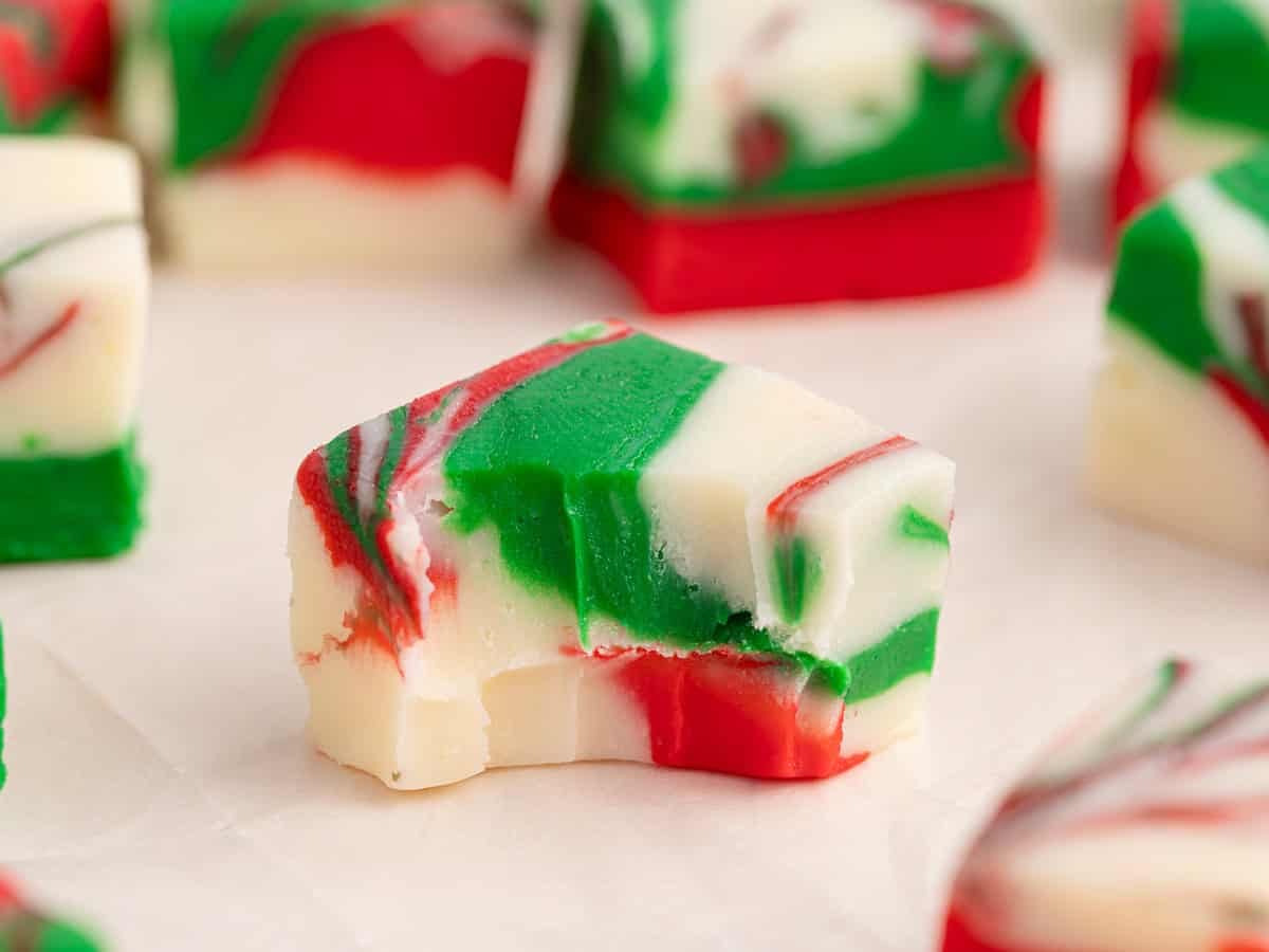 A piece of red, green, and white swirled Christmas Fudge with a bite taken out sits on parchment paper, with more pieces in the background, showcasing the festive treat from a classic Christmas Fudge Recipe.