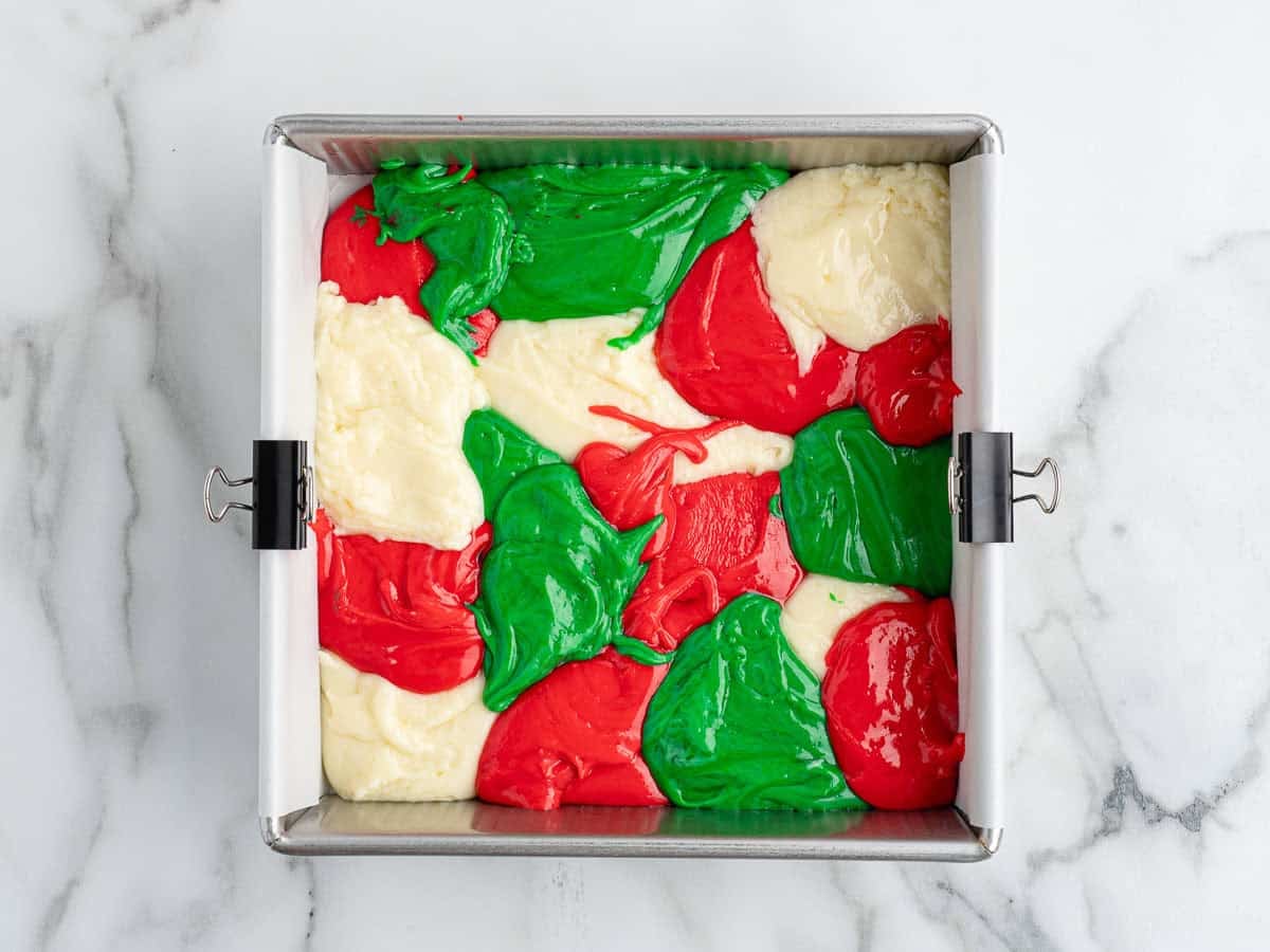 A square baking pan with blobs of red, green, and white fudge mixture, arranged in a festive swirl pattern reminiscent of a Christmas Fudge Recipe, sits on a marble countertop.