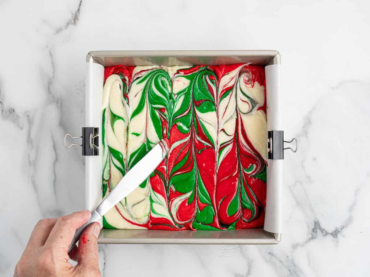 A hand spreads a marbled red, green, and white fudge mixture in a baking pan lined with parchment paper, creating a festive swirl pattern on a white marble surface, perfect for making your favorite Christmas Fudge recipe.