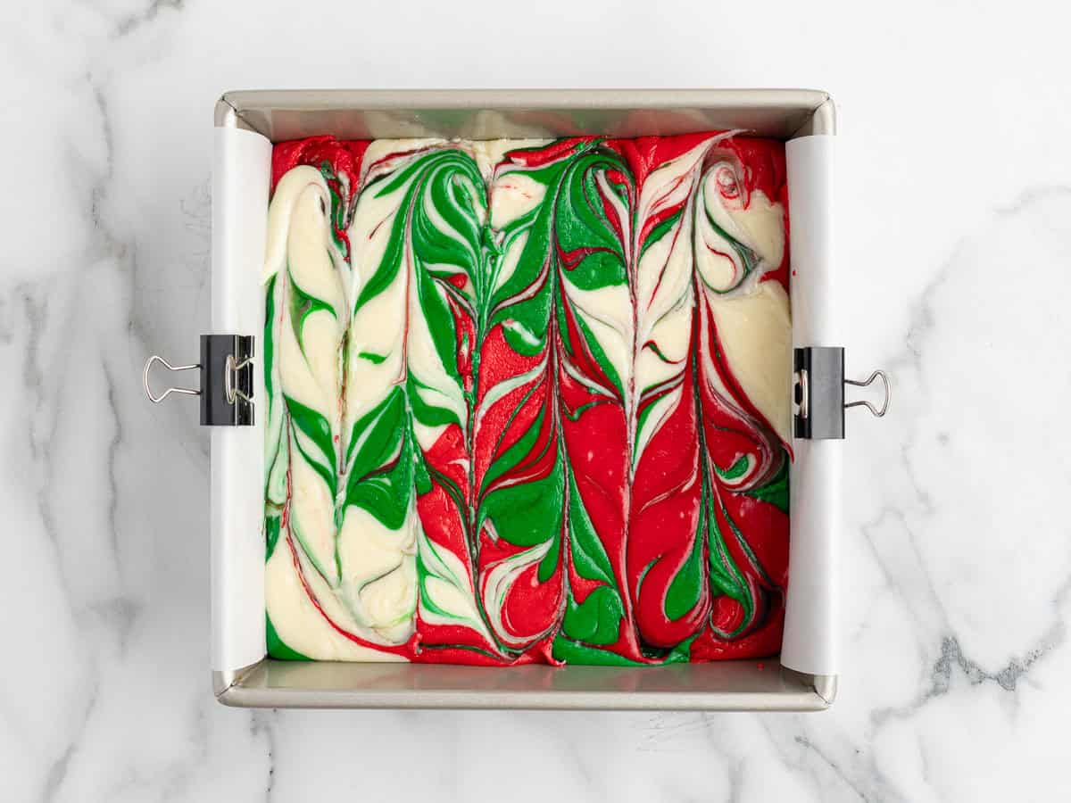 A square baking pan with swirled red, green, and white fudge, creating a festive marbled pattern perfect for a Christmas Fudge recipe. The pan is lined with parchment paper and held with metal clips on each side.
