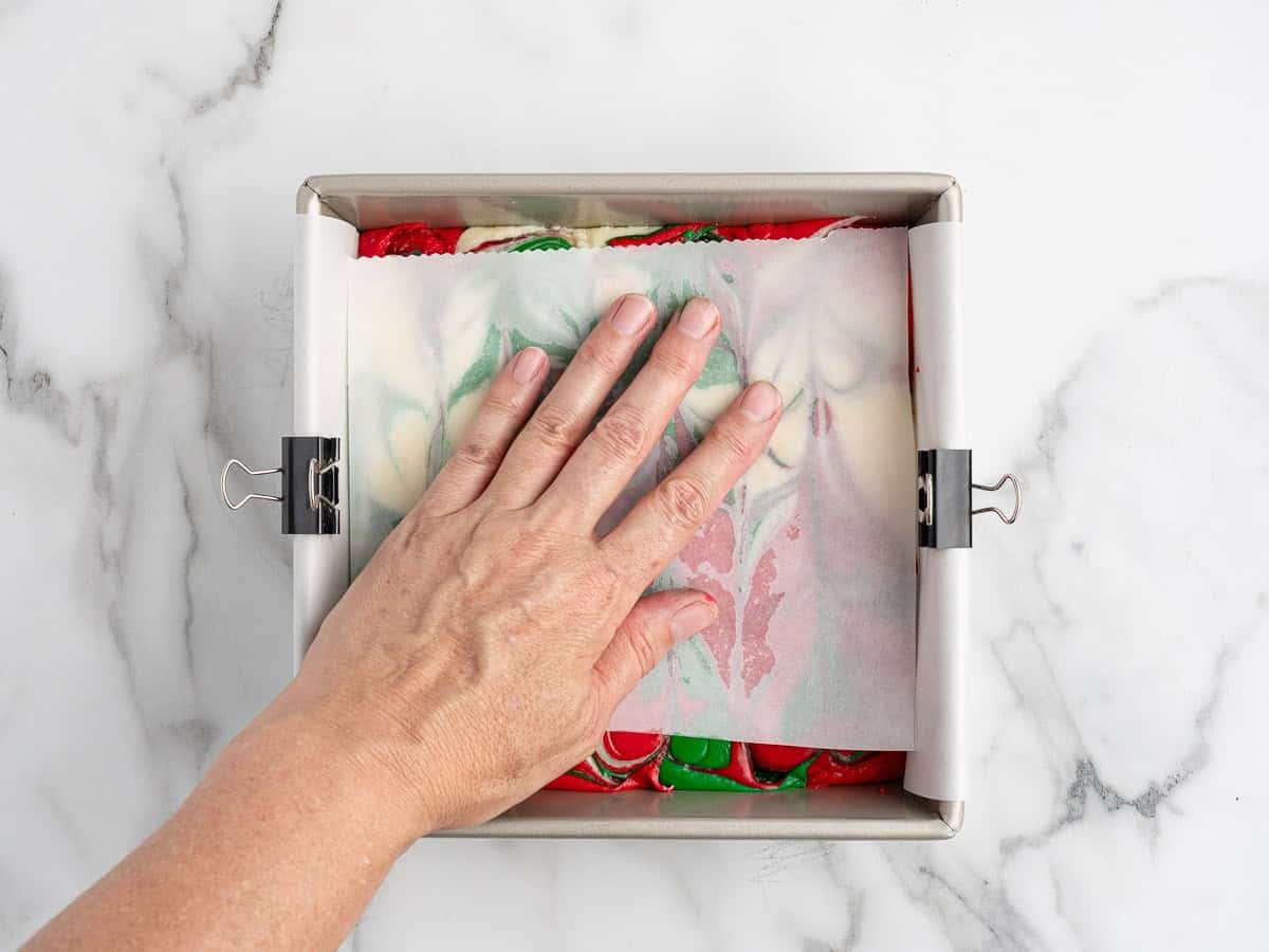 A hand presses down on a square pan lined with parchment paper, filled with vibrant red, green, and white swirled Christmas Fudge. The parchment is held in place with metal binder clips on both sides, showcasing the festive results of a classic Christmas Fudge Recipe.
