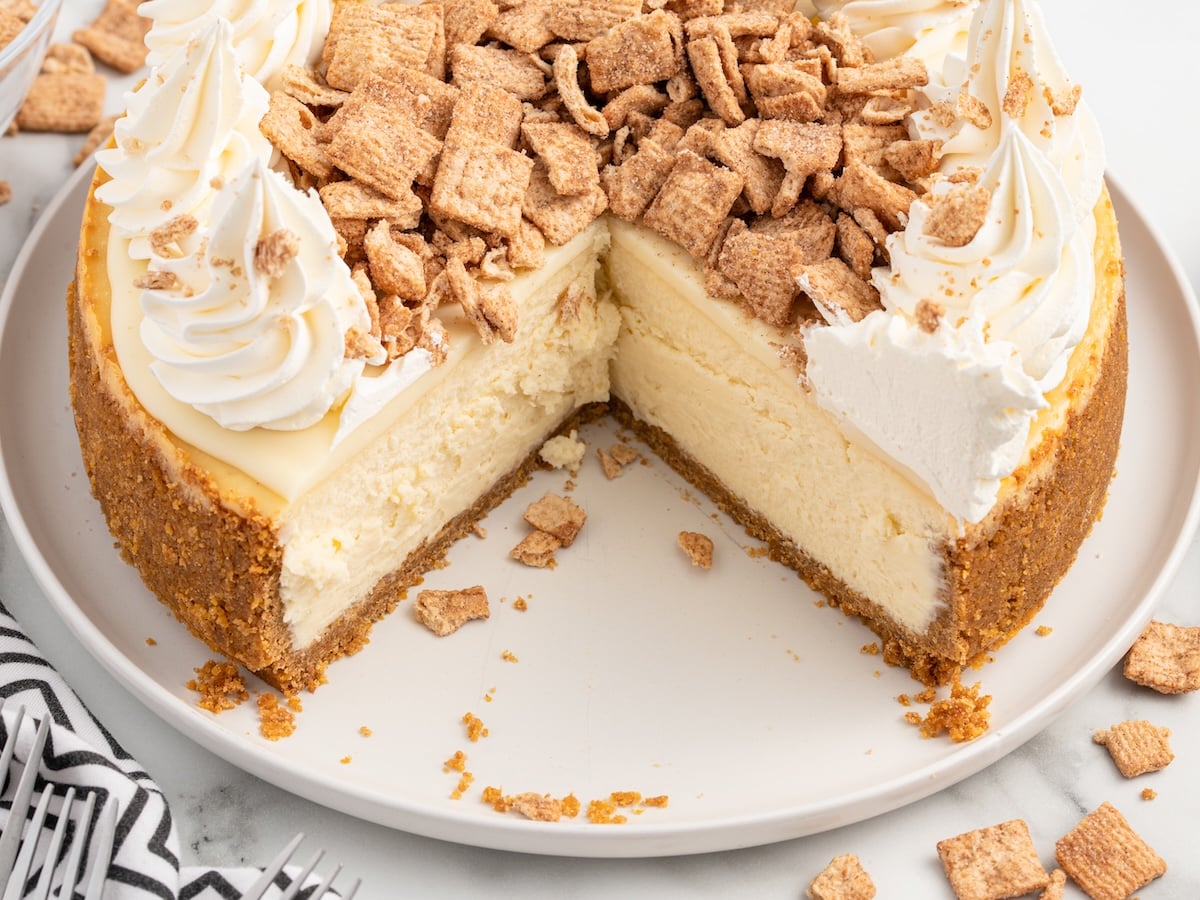 Cinnamon Toast Crunch cheesecake with a slice removed showing the creamy filling, cereal crust, whipped cream rosettes, and crushed cereal topping.