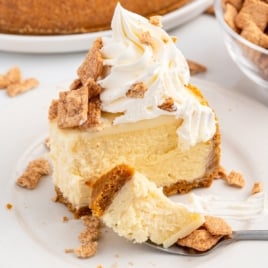A slice of creamy Cinnamon Toast Crunch Cheesecake topped with whipped cream and cinnamon cereal pieces sits on a white plate, with a fork holding a bite-sized piece in the foreground. A bowl of cereal is visible in the background.
