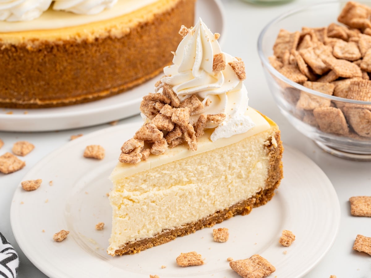A slice of Cinnamon Toast Crunch Cheesecake with a graham cracker crust is topped with whipped cream and crunchy cinnamon cereal pieces, served on a white plate next to a bowl of cereal.