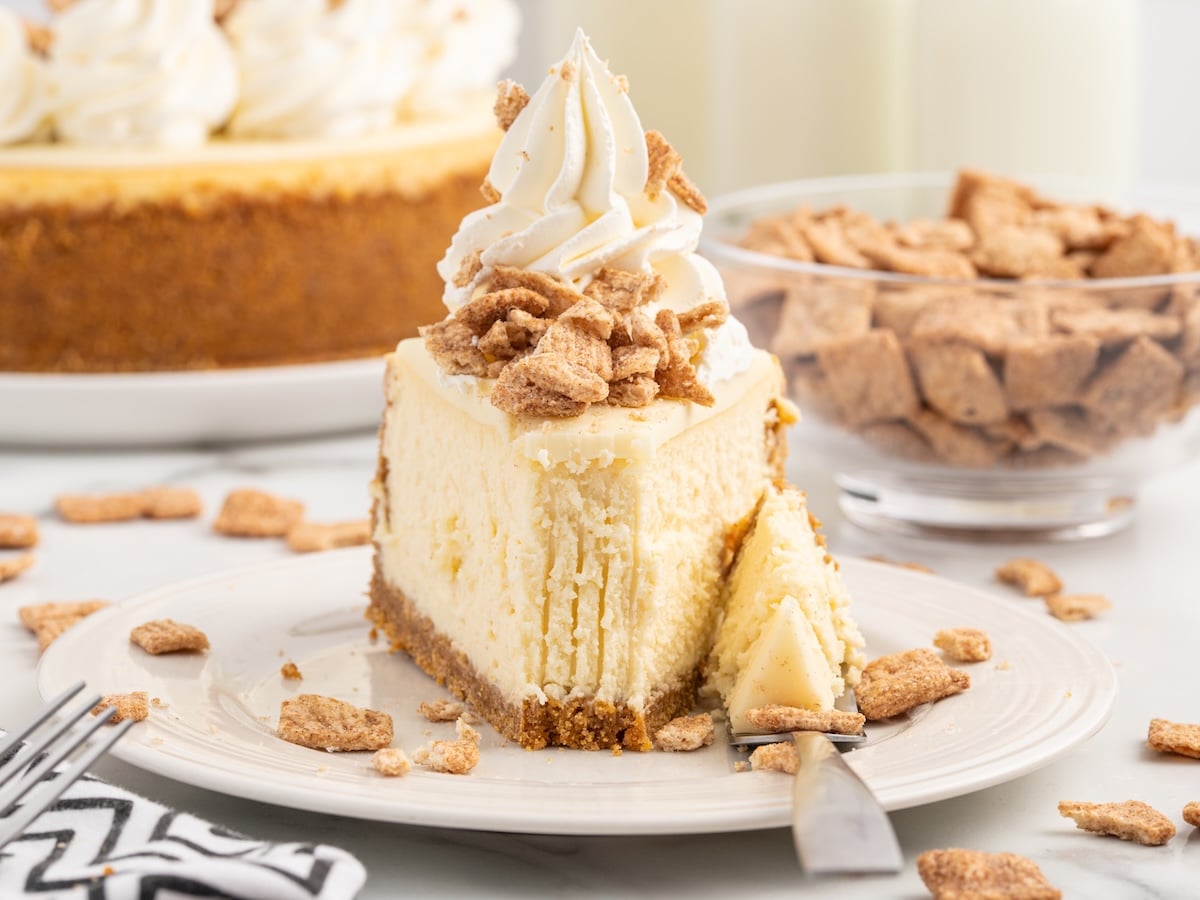 A slice of Cinnamon Toast Crunch Cheesecake topped with whipped cream and crunchy cinnamon cereal sits on a white plate, with a fork and a bite cut out. A bowl of cereal and the rest of the cheesecake are in the background.