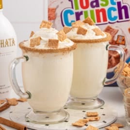 Two creamy drinks in clear mugs with whipped cream, cinnamon sugar rims, and Cinnamon Toast Crunch cereal pieces on top. A box of Toast Crunch cereal, bottle of horchata liqueur, and loose cereal are in the background—everything you need to craft a delicious Cinnamon Toast Crunch Cocktail.