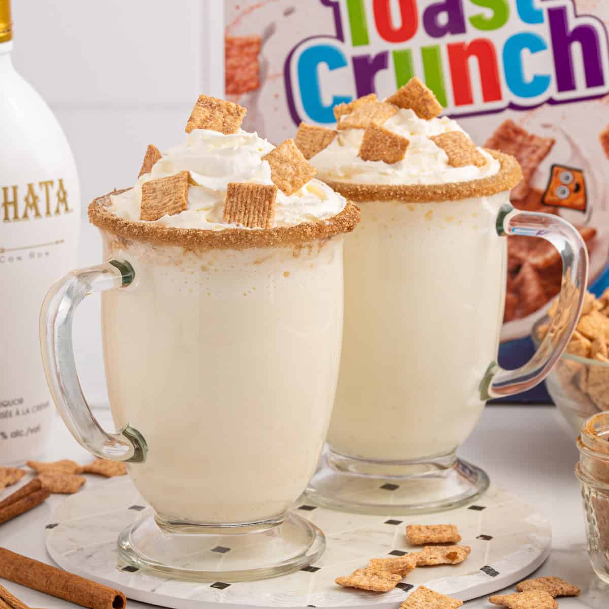 Two creamy drinks in clear mugs with whipped cream, cinnamon sugar rims, and Cinnamon Toast Crunch cereal pieces on top. A box of Toast Crunch cereal, bottle of horchata liqueur, and loose cereal are in the background—everything you need to craft a delicious Cinnamon Toast Crunch Cocktail.