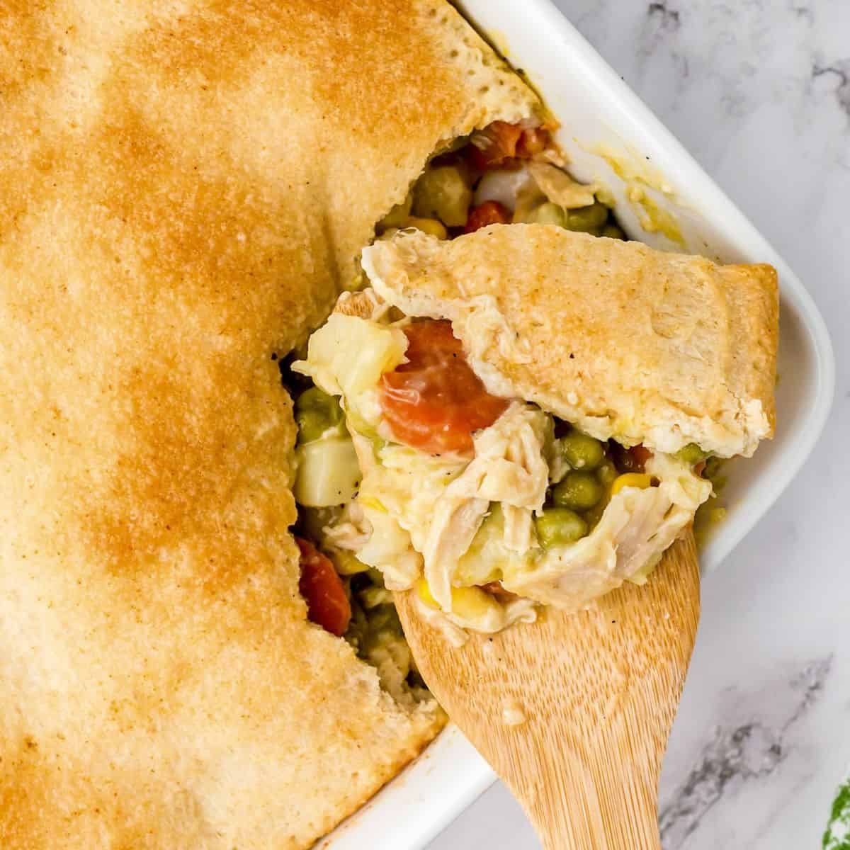 A golden-brown Easy Chicken Pot Pie with a flaky crust is being served with a wooden spoon, revealing a hearty filling of chicken, peas, carrots, and other vegetables.