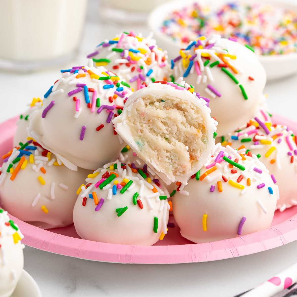 Plate of no bake cheesecake truffles coated in white chocolate and topped with colorful sprinkles, with one truffle cut open to show creamy funfetti filling