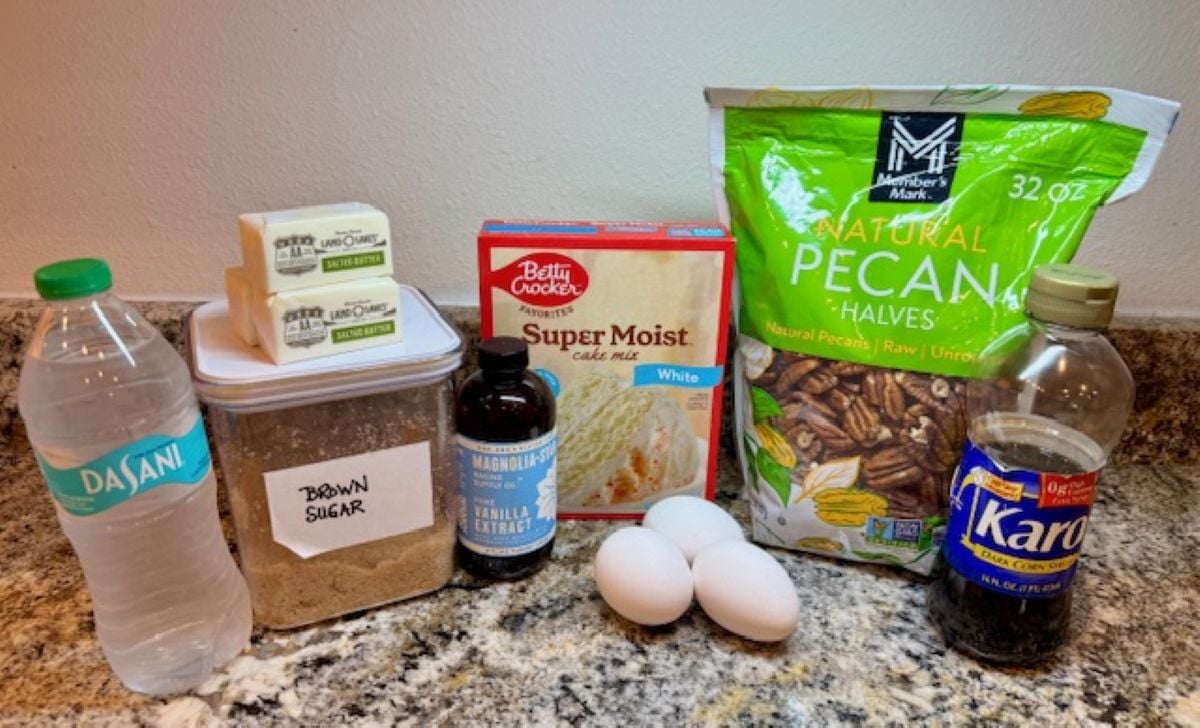 A variety of baking ingredients on a counter for Pecan Dump Cake, including a bottle of water, brown sugar, vanilla extract, three eggs, white cake mix, a stick of butter, pecan halves, and corn syrup.