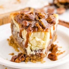 A slice of decadent Pecan Pie Cheesecake with a graham cracker crust, topped with caramel sauce and pecans, sits on a white plate—bringing the flavors of your favorite Pecan Pie Cheesecake Recipe to life.