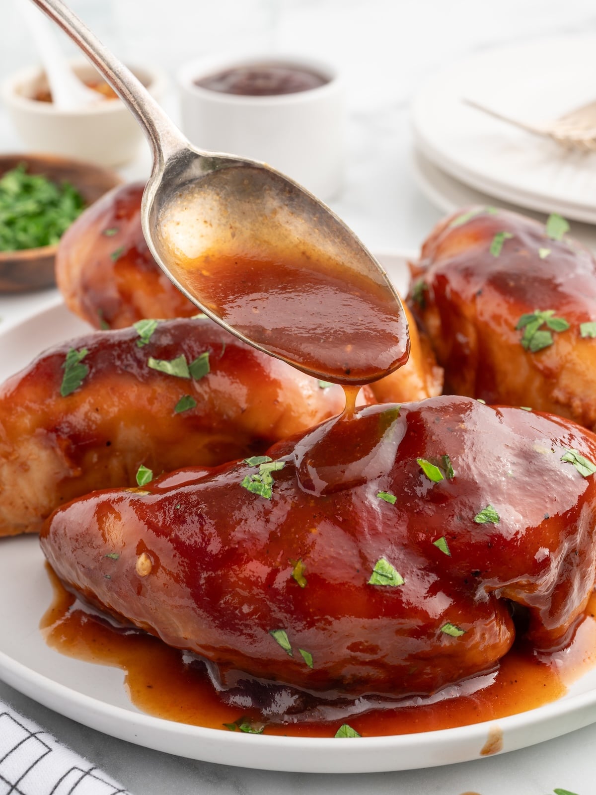 A spoon drizzles barbecue sauce over glazed Sweet Baby Rays Crockpot Chicken breasts on a white plate, garnished with chopped parsley. In the background, there are small bowls, a fork, and a white napkin.