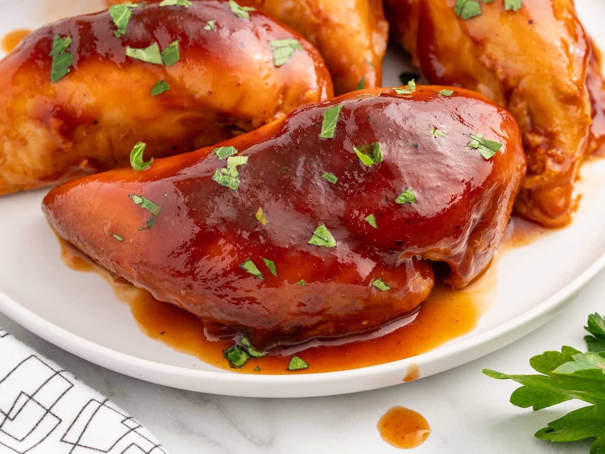 Close-up of glazed Sweet Baby Rays Crockpot Chicken breasts garnished with chopped parsley on a white plate, with sauce pooling around the chicken. A white napkin and some parsley are visible in the corner.