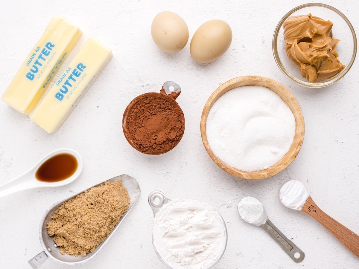 An overhead view of baking ingredients on a white surface, ready to make Chocolate Peanut Butter Cookies, including butter, eggs, peanut butter, cocoa powder, sugar, flour, brown sugar, vanilla extract, and baking soda.