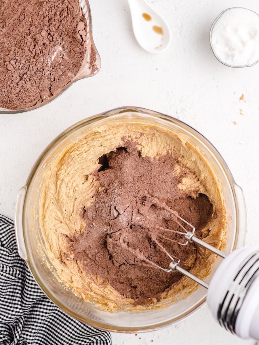 A close-up shot shows cocoa powder being added to a bowl of creamy batter for Chocolate Peanut Butter Cookies, while a white electric mixer with beaters is mixing it.