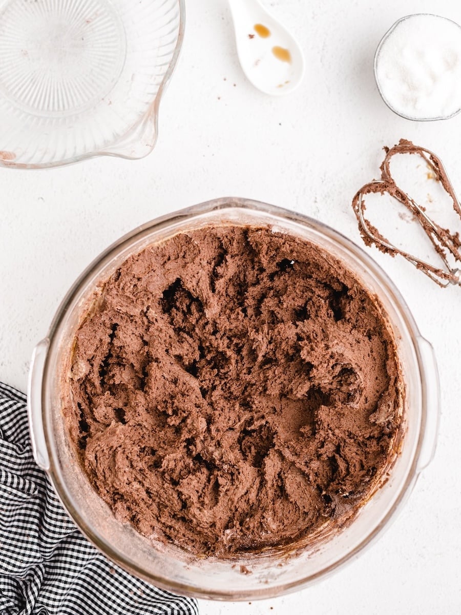A bowl of rich, chocolate cake batter sits on a white surface, ready to become luscious Chocolate Peanut Butter Cookies, with a measuring cup, a spoon of vanilla extract, and a small bowl of sugar nearby.