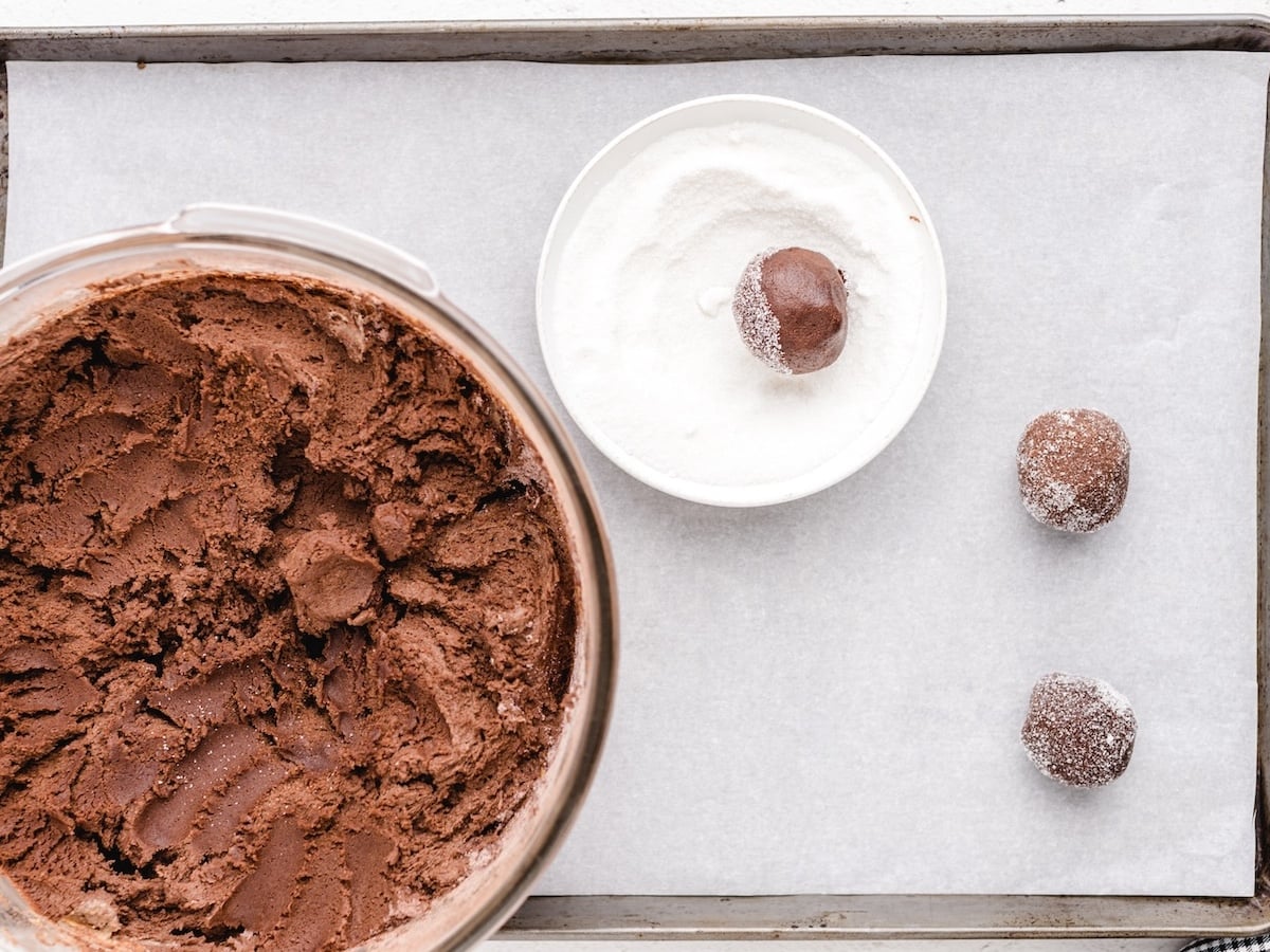 A tray with chocolate cookie dough in a bowl, a small bowl of sugar holding a dough ball, and two sugar-coated dough balls on parchment paper, ready for baking these delicious Chocolate Peanut Butter Cookies.