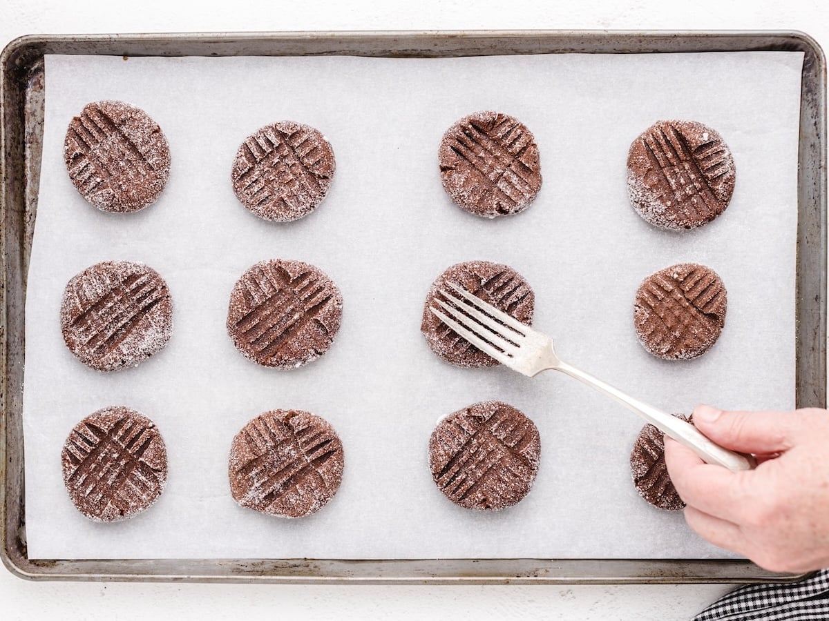 Chocolate Peanut Butter Cookies are coated in sugar and arranged on a baking sheet lined with parchment paper. A fork is used to press a crisscross pattern onto one of the cookies, making this Chocolate Peanut Butter Cookies Recipe extra special.
