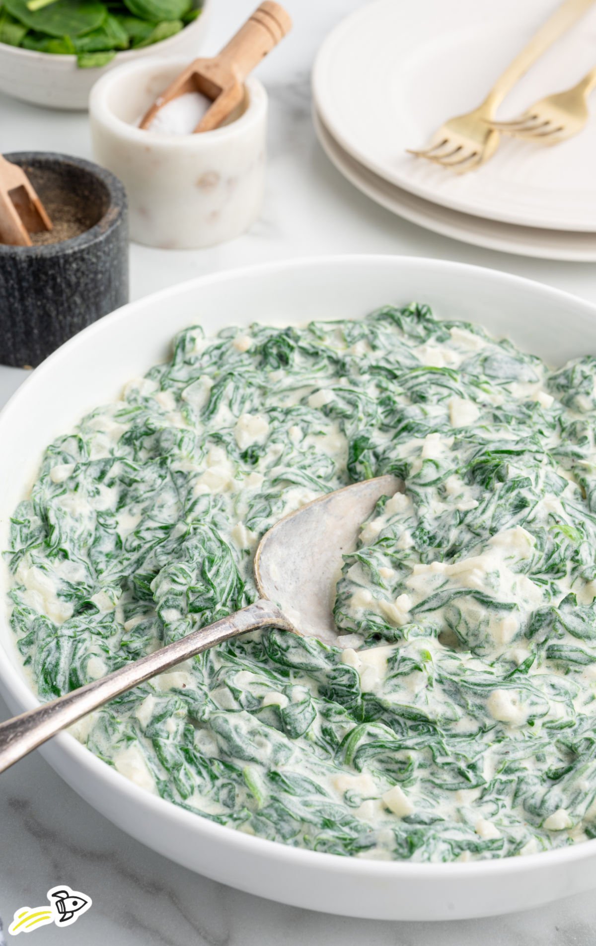 Bowl of homemade creamed spinach with a spoon, showing the rich, creamy sauce coating tender spinach leaves. Salt and pepper containers and plates in the background create a cozy, homemade feel.