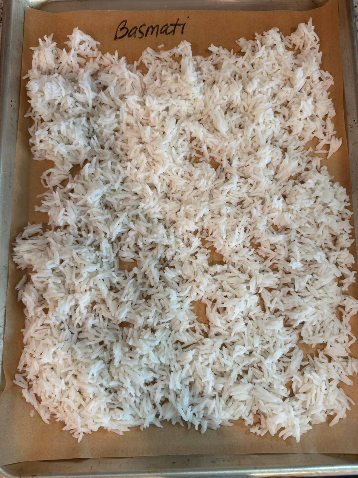 Cooked basmati rice is spread in a single layer on a parchment-lined baking sheetโa perfect prep step for any Better Than Take-Out Fried Rice Recipe. The word Basmati is written at the top of the parchment.