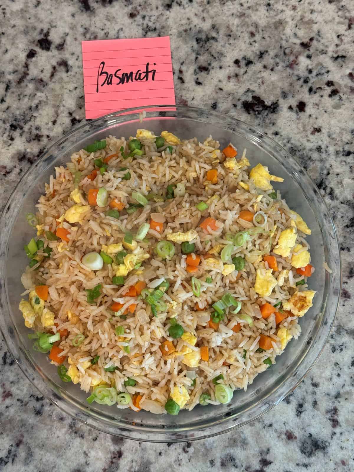A glass bowl filled with Better Than Take-Out Fried Riceโscrambled eggs, chopped green onions, carrots, and peasโrests on a granite countertop with a pink sticky note labeled Basmati beside it.