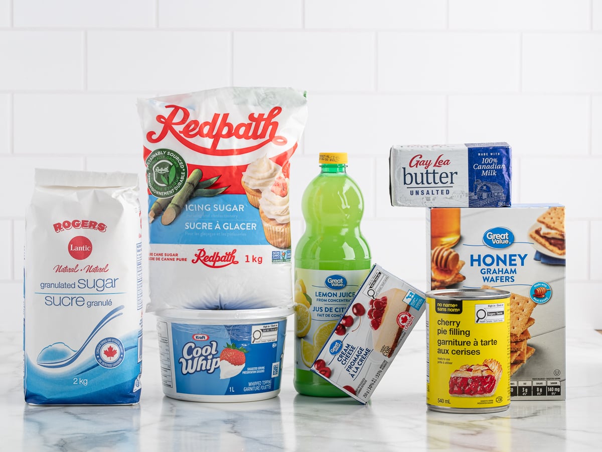 Assorted baking ingredients on a marble countertop, including those needed for No Bake Cherry Cheesecake&mdash;granulated sugar, icing sugar, lemon juice, whipped topping, butter, canned cherry pie filling, condensed milk, and honey graham wafers.