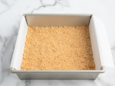 A square metal baking pan lined with parchment paper, filled with an even layer of graham cracker crumb crust for No Bake Cherry Cheesecake, sits on a white marble surface.