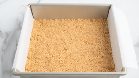 A square metal baking pan lined with parchment paper, filled with an even layer of graham cracker crumb crust for No Bake Cherry Cheesecake, sits on a white marble surface.
