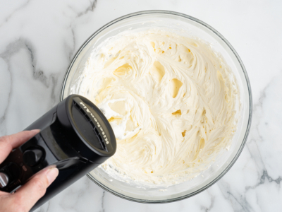 A hand holds a black electric mixer over a glass bowl filled with creamy, whipped mixture—perfect for creating a No Bake Cherry Cheesecake—on a white marble surface.