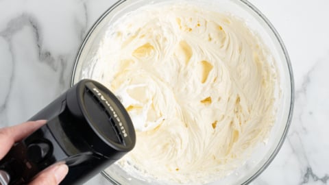 A hand holds a black electric mixer over a glass bowl filled with creamy, whipped mixture&mdash;perfect for creating a No Bake Cherry Cheesecake&mdash;on a white marble surface.