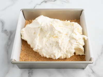 A square baking pan lined with parchment paper holds a graham cracker crust and a generous dollop of creamy cheesecake filling—perfect for creating a delicious No Bake Cherry Cheesecake.