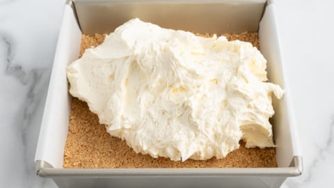 A square baking pan lined with parchment paper holds a graham cracker crust and a generous dollop of creamy cheesecake filling&mdash;perfect for creating a delicious No Bake Cherry Cheesecake.