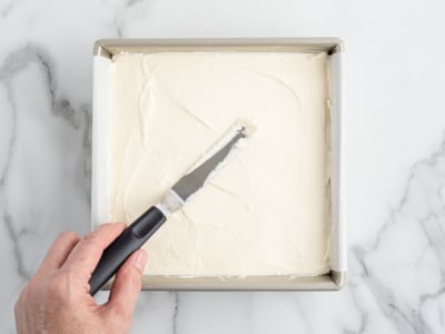 A hand uses an offset spatula to spread creamy white frosting evenly over a square No Bake Cherry Cheesecake in a metal baking pan on a marble surface.