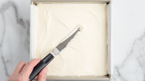 A hand uses an offset spatula to spread creamy white frosting evenly over a square No Bake Cherry Cheesecake in a metal baking pan on a marble surface.