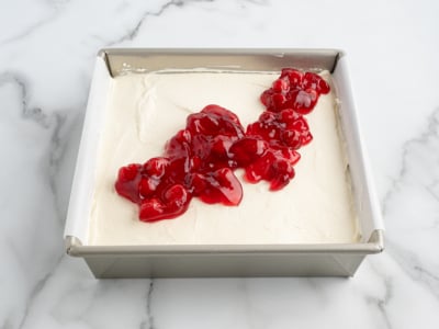 A square baking pan filled with a creamy white No Bake Cherry Cheesecake mixture, topped with a strip of glossy red cherry pie filling, sits on a white marble surface.