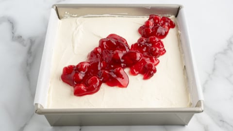 A square baking pan filled with a creamy white No Bake Cherry Cheesecake mixture, topped with a strip of glossy red cherry pie filling, sits on a white marble surface.
