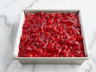A square metal baking pan lined with parchment paper is filled with bright red cherry pie filling, perfect for a No Bake Cherry Cheesecake, with whole cherries and glossy syrup set on a white marble surface.
