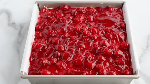 A square metal baking pan lined with parchment paper is filled with bright red cherry pie filling, perfect for a No Bake Cherry Cheesecake, with whole cherries and glossy syrup set on a white marble surface.